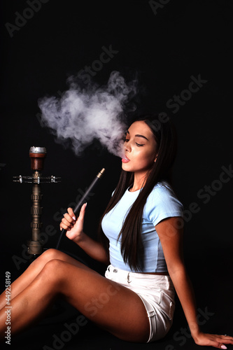 The girl smokes a hookah on a black background. Smoke comes out of the mouth. The pleasure of smoking