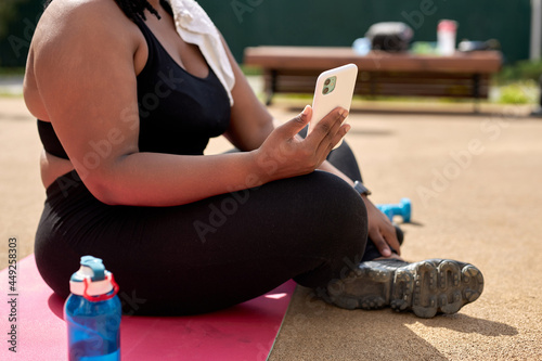 Cropped Photo Of Fat African Woman Sitting On Fitness Mat Using Smartphone, Tired After Sport Exercises, Take A Break During Workout, Alone Outdoors In Sports Ground. Side View Portrait