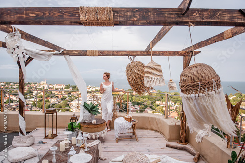 Young fashion woman in white jumpsuit with styling, holding glass of white wine, stands on the open terrace of the roof of high house overlooking the sea. Stylish girl in Moroccan interior celebrating