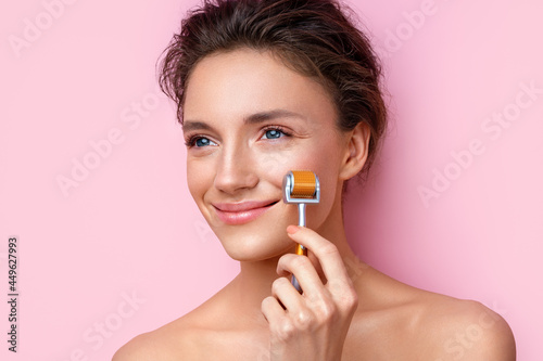 Beautiful woman using derma roller for her facial skin. Photo of woman on pink background. Beauty and skin care concept