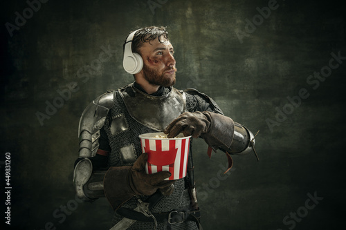 Portrait of one brutal bearded man, medeival warrior or knight in headphones with bucket of popcorn over dark background.