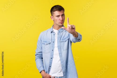 Lifestyle, people emotions and summer leisure concept. Serious-looking displeased blond man shaking finger as scolding person, give warning or prohibit bad action, disagree over yellow background