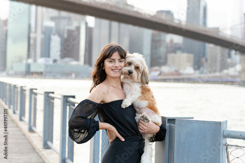 a woman holding her small mixed breed dog at the boardwalk by the hudson river with brooklyn bridge and new york city buildings in the background