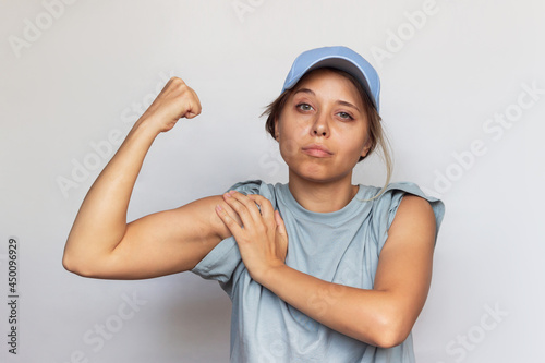 Strong confident caucasian young blonde woman in a gray t-shirt and cap raises arm and shows bicep isolated on a white background. Feminism, girl power, equal women's rights, independence concept