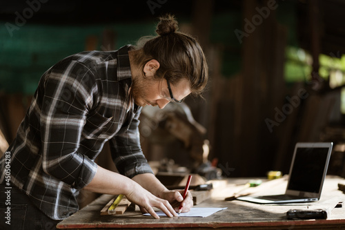 young hipster carpenter man working with computer laptop in workshop . craftsman wearing safety glasses checking order of clients or learning online