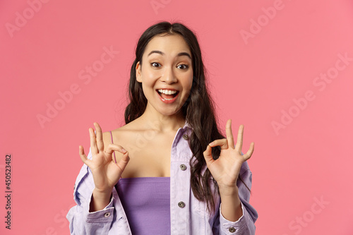 Excellent choice. Close-up portrait of happy amused young stylish asian girl satisfied, showing okay signs and smiling, nod in approval, totally agree, think product super cool, pink background
