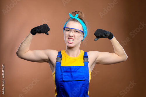 Portrait of a strong attractive Caucasian young woman in construction worker workwear and safety goggles. Demonstrates his strength by showing biceps and making a grimace. Looks into the camera