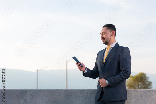Man buying assets from his mobile device. Young man using his smartphone to make investments. Young executive in blue suit standing at the entrance of his company, checking the contact list. 
