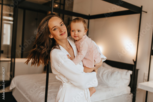 Portrait of positive mother and little daughter in home outfit posing in bedroom. Woman in bathrobe hugging baby