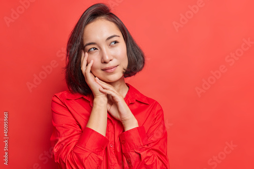Dreamy young Asian woman with dark hair keeps hands near face has thoughtful expression imagines something in mind wears red shirt in one tone with background. Monochrome shot. Let me think.