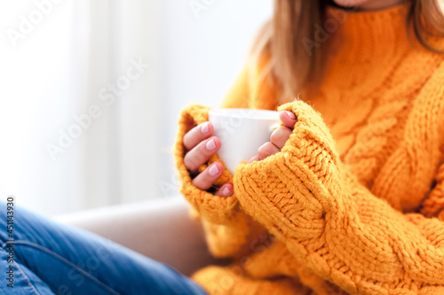 Cup of coffee in female hands wearing in knitted cozy sweater with long warm sleeves. Woman drinking calming drink at home.