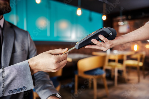 Hands holding a payment terminal. A man elegantly dressed in a business suit puts a credit or debit card in an online payment terminal held by a feminine hand. Paying bills for a meal at a restaurant