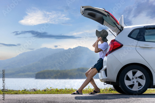 Young Asian woman traveler sitting on hatchback car trunk with mountain landscape background. Traveler car concept