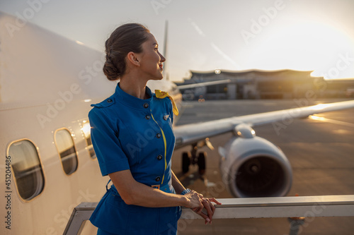 Thoughtful air stewardess in blue uniform looking away, standing outdoors at the sunset. Commercial airplane near terminal in an airport in the background. Aircrew, occupation concept