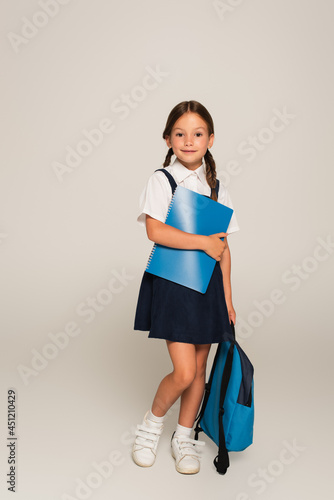 full length view of smiling schoolgirl standing with blue backpack and copy books on grey