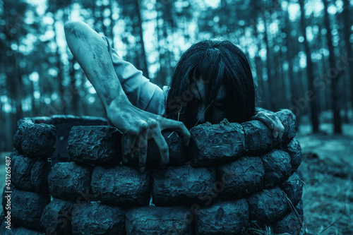Girl in image of scary zombie crawls out of the stone well in dark forest.