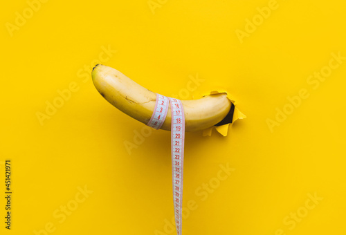 A banana with a tape measure wrapped around it appears through a torn hole in yellow paper. The concept of healthy food, diet and potency. Copy space.