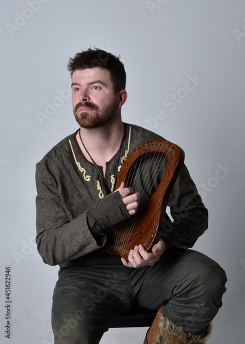 Full length portrait of young handsome man wearing medieval Celtic adventurer costume playing a small musical harp, isolated on studio background.