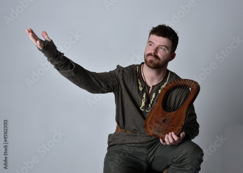 Full length portrait of young handsome man wearing medieval Celtic adventurer costume playing a small musical harp, isolated on studio background.