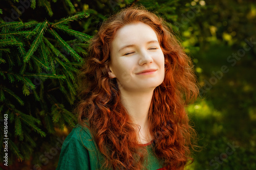 A large portrait of a beautiful woman with long red hair against the background of greenery in the park. Her eyes are peacefully closed, the woman enjoys nature.