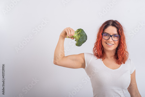 Caucasian woman holding broccoli and demonstrating biceps over white background.