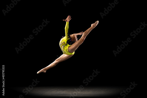 Slim sportive girl, rhythmic gymnastics artist in bright stage costume isolated on dark studio background in ray of light. Concept of sport, action, aspiration, active lifestyle