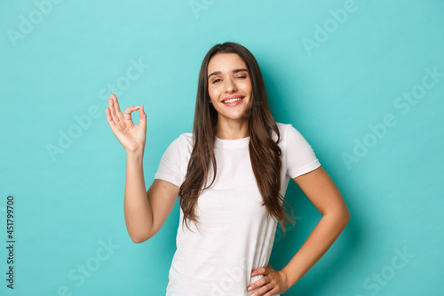 Image of confident smiling woman in white t-shirt, showing okay sign and looking pleased, agree or approve something good, like and recommend product, standing over blue background