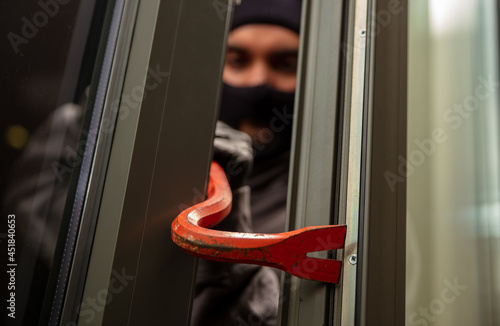 Thief in balaclava holding a crowbar to break a glass window.