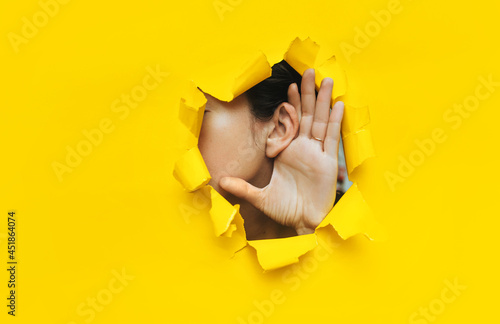 Close-up of a woman's ear and hand through a torn hole in the paper. Yellow background, copy space. The concept of eavesdropping, espionage, gossip and tabloids.