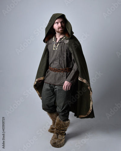 Full length portrait of young handsome man wearing medieval Celtic adventurer costume with hooded fur cloak, standing pose isolated on studio background.