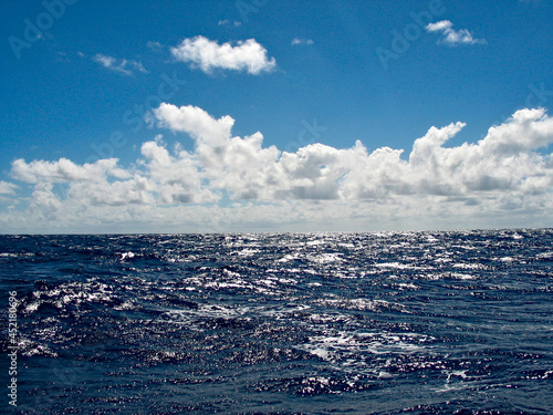 View from the deck of a sailboat crossing an ocean, midday hard sun, trade wind cumulus clouds.