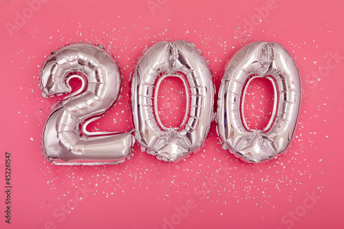 From above of silver shiny balloons demonstrating number 200 on pink background with scattered glitter
