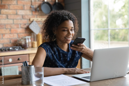 Happy millennial African woman recording, sending audio message on smartphone, using voice recognition app. Remote employee, student giving command to virtual assistant on mobile phone at laptop