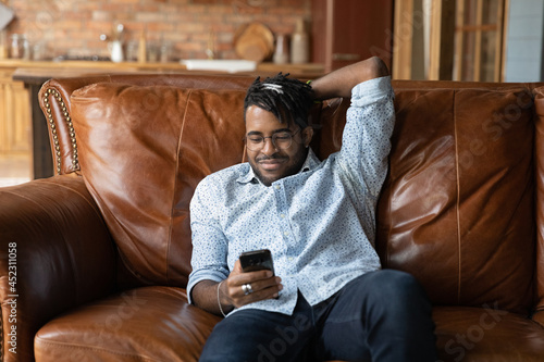 Happy lazy African American guy using smartphone, enjoying leisure at home, relaxing on leather couch, overusing mobile phone, texting message, chatting online. Cellphone addict, social media obsess