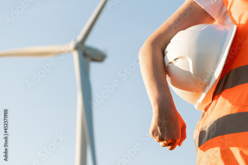 An engineer with a helmet in his hands and orange vesta stands on the background of windmill or wind turbine with copy space.