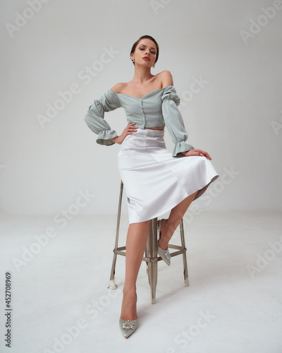 Elegant woman in pastel green top, silver skirt and high heels sitting at high metal chair with hand at waist and long legs. Stunning gorgeous fashion female model with natural make-up and pony tail