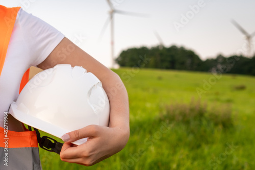 An engineer with a helmet in his hands and orange vesta stands on the background of windmill or wind turbine with copy space.