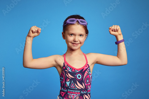 Little girl spends leisure time in water park, raises arms and shows muscles, ready for swim, poses over blue background, wears goggles and swimsuit