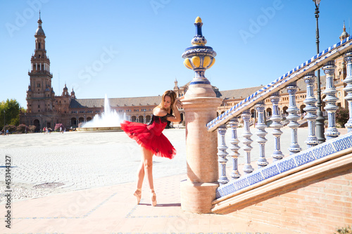 ballet dancer with red tutu leaning on a park railing in seville. The dancer makes different postures and stretches on the railing. Classical ballet concept.
