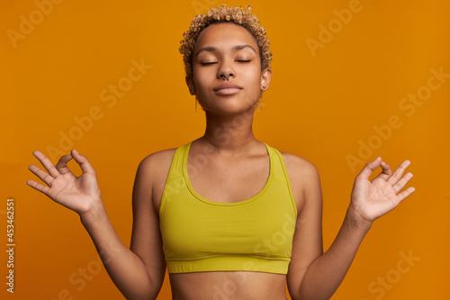 Portrait of relaxed, meditating African woman reaching zen and inner harmony, making Om sign with fingers, looking peaceful, feeling balanced enjoying spiritual practice isolated on orange wall