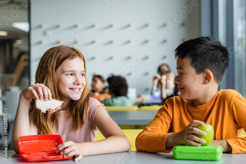 redhead girl with sandwich smiling to asian boy in school dining room