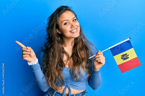 Young hispanic girl holding moldova flag smiling happy pointing with hand and finger to the side