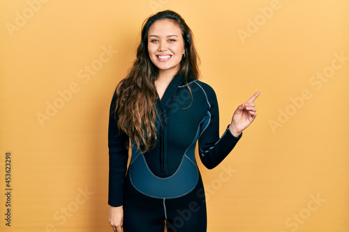 Young hispanic girl wearing diver neoprene uniform with a big smile on face, pointing with hand finger to the side looking at the camera.