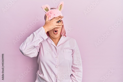 Hispanic woman with pink hair wearing sleep mask and pajama peeking in shock covering face and eyes with hand, looking through fingers with embarrassed expression.