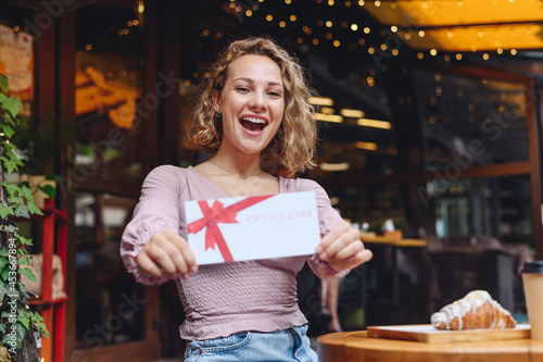 Young happy smiling woman in casual clothes sitting alone at table in coffee shop cafe eat breakfast hold gift certificate coupon voucher card for store relaxing in restaurant during free time indoor
