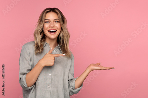 Excited young woman holding palm open for new product or sale presentation. Overjoyed happy blond girl with open mouth point finger at arm with copy space for promotion isolated over pink studio wall