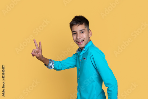 Young smiling teenage boy gestures a sign victory or pease against a yellow background.