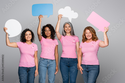 Smiling multiethnic women with pink ribbons holding speech bubbles on grey background
