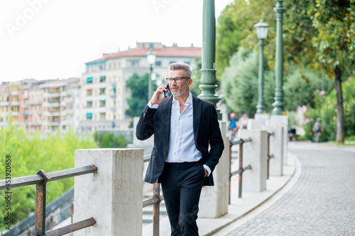 Businessman in good mood is talking on cellphone on his way to office. Handsome cheerful manager in formal suit walking enjoying getting to job in morning by foot having mobile conversation