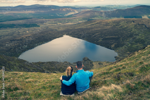 Couple sitting on the top of the mountain. View from behind.. Heart shaped Lake Ouler Tonelagee Mountain, Wicklow County, Ireland.
